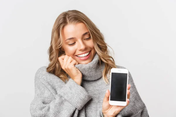Närbild av härlig blond flicka ser glad på mobiltelefon skärm, står i grå tröja över vit bakgrund — Stockfoto