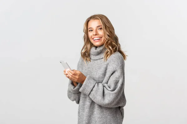 Porträtt av attraktiv glad kvinna i grå tröja, håller smartphone och ler mot kameran, begreppet online shopping och sociala nätverk — Stockfoto