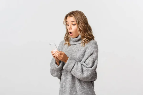 Porträtt av förvånad blond flicka ser upphetsad på smartphone skärm, står över vit bakgrund. Begreppet teknik och kommunikation — Stockfoto