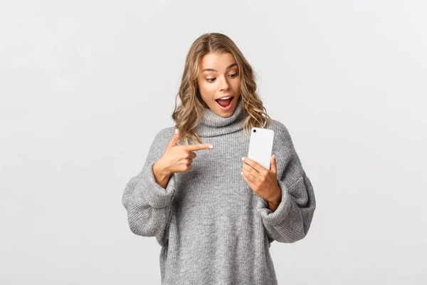 Porträtt av attraktiva kaukasiska kvinnliga modellen i grå tröja, ser förvånad och pekar på mobiltelefon, rekommendera något, vit bakgrund — Stockfoto