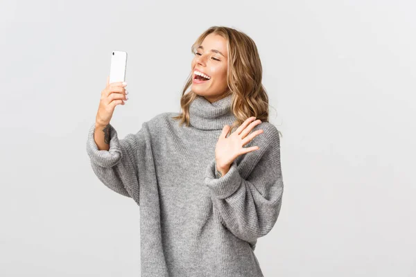 Porträtt av attraktiv kaukasiska flicka i grå tröja, hålla mobiltelefon och vinka åt kameran för att säga hej, med videosamtal, står över vit bakgrund — Stockfoto
