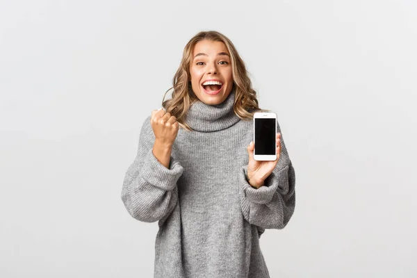 Bild av vinnande glad blond flicka, triumferande över online prestation, visar smartphone skärm och göra knytnäven pump, vit bakgrund — Stockfoto