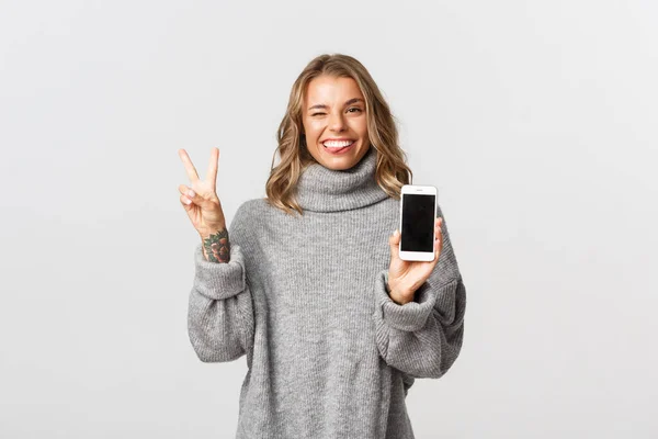 Porträtt av attraktiv blond flicka med tatuering, visar smartphone skärm och fred tecken, stick tunga och leende glad, står över vit bakgrund — Stockfoto