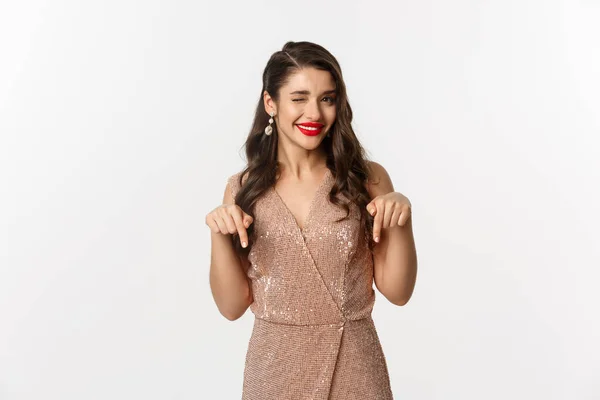 Elegantní ženský model s červenými rty a náušnicemi, ukazující prsty na vánoční nabídku, mrkající a usmívající se, ukazující promo, stojící ve společenských šatech na bílém pozadí — Stock fotografie