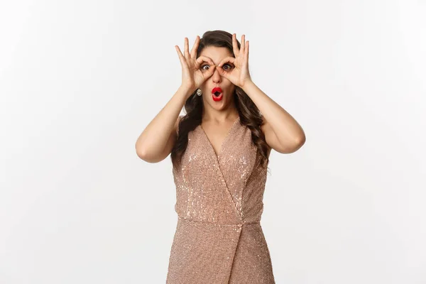 Fiesta de Navidad y concepto de celebración. Mujer en vestido de glamour y labios rojos mirando algo increíble, mirando a través de los prismáticos de mano, de pie sobre fondo blanco — Foto de Stock