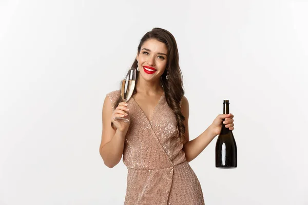 Концепция зимних праздников. Изображение счастливой молодой женщины в роскошном платье, пьющей шампанское из стекла, держа бутылку, новогодняя вечеринка, белый фон — стоковое фото