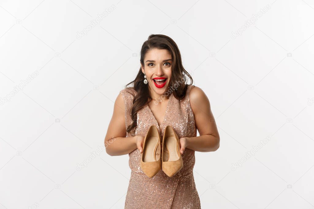 Party and celebration concept. Excited woman showing new pair of heels and smiling, wearing elegant dress, standing over white background
