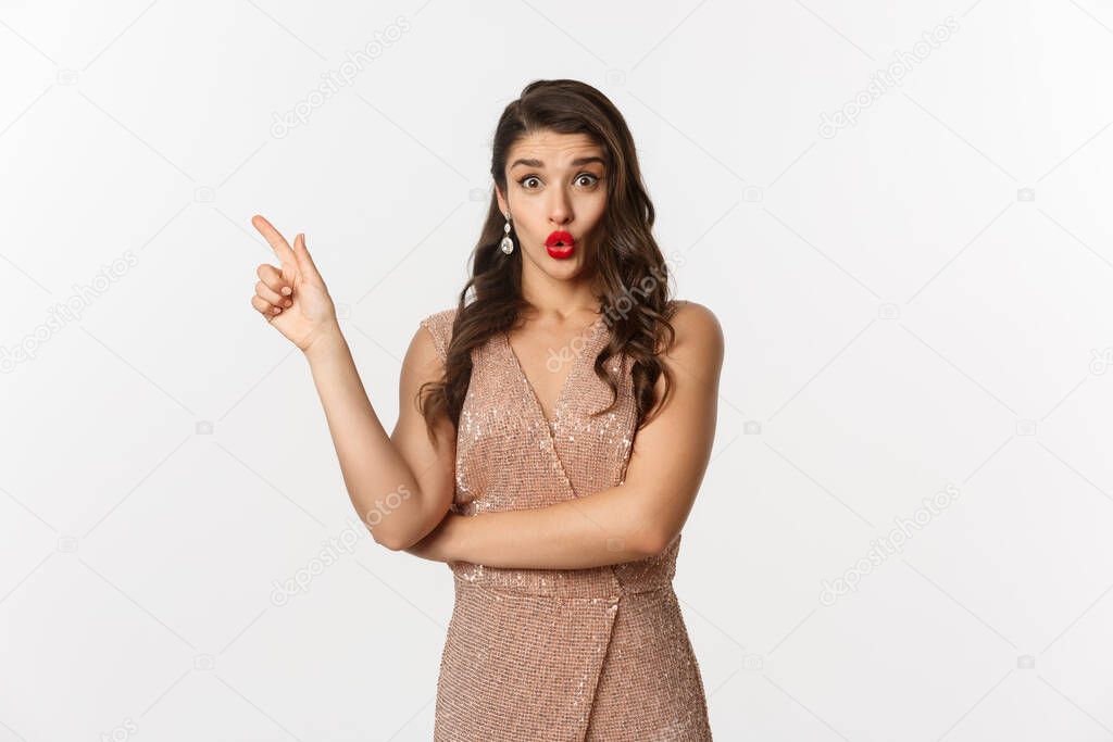 Christmas, holidays and celebration concept. Intrigued beautiful woman asking question about promo offer, pointing finger left at logo or banner, standing in party dress