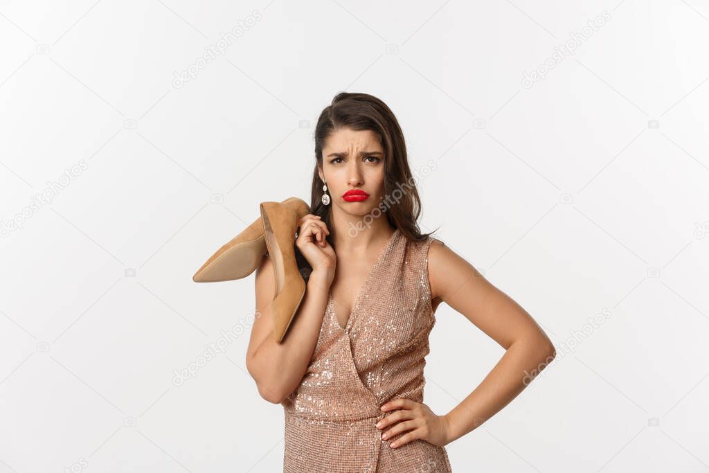 Party and celebration concept. Image of attractive clingy woman complaining, tired feet after wearing heels, holding shoes in hands and sulking, standing over white background