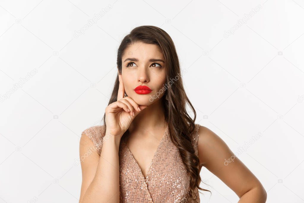 Concept of New Year celebration and winter holidays. Close-up of elegant woman with red lips and dress, looking at upper left corner and thinking, standing over white background