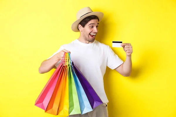 Hombre excitado yendo de compras de vacaciones, mirando satisfecho en la tarjeta de crédito, sosteniendo bolsas de papel, de pie sobre el fondo amarillo —  Fotos de Stock