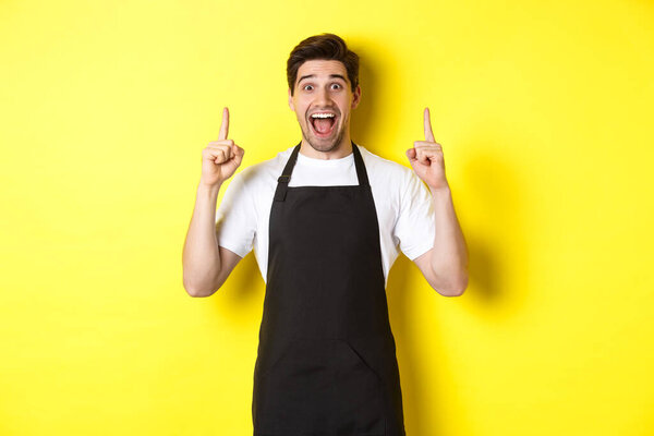 Excited coffee shop owner in black apron pointing fingers up, showing special offers, standing over yellow background