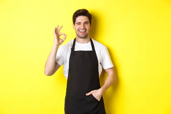 Уверенный и красивый официант показывает знак ОК, в черном фартуке и стоя на желтом фоне — стоковое фото