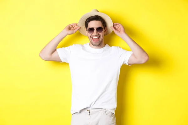 黄色の背景に立っている間、笑顔、休暇中の幸せな男は、わら帽子とサングラスを身に着けている — ストック写真
