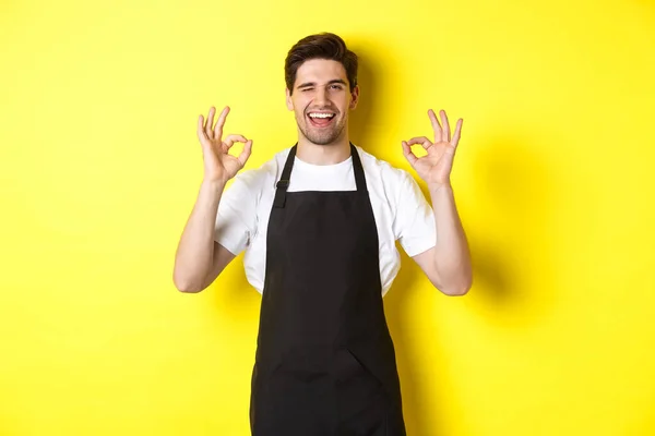 Jovem barista masculino em avental preto mostrando OK sinais e sorrindo, garantia de qualidade no café, de pé sobre fundo amarelo — Fotografia de Stock