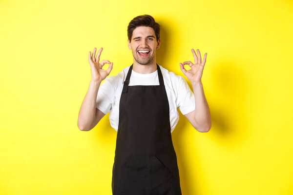 Young male barista in black apron showing OK signs and smiling, guarantee quality in coffee shop, standing over yellow background