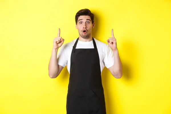Здивований чоловічий офіціант у чорному фартусі, який показує логотип або рекламу, вказуючи пальці вгору на копіювальному просторі, стоячи над жовтим тлом — стокове фото