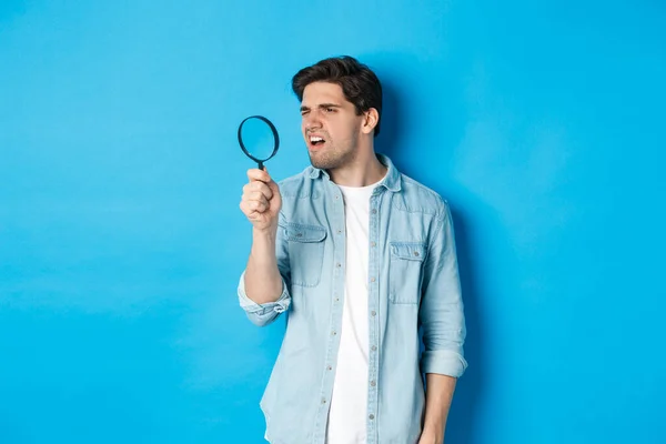 Jeune homme réfléchi regardant à travers la loupe, lire quelque chose de minuscule, debout sur fond bleu — Photo