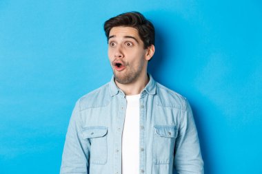 Şaşırmış, soldan etkilenmiş, ağzı açık, büyüleyici, sıradan kıyafetler giymiş, mavi arka planda duran yakışıklı adamın yakın çekimi.