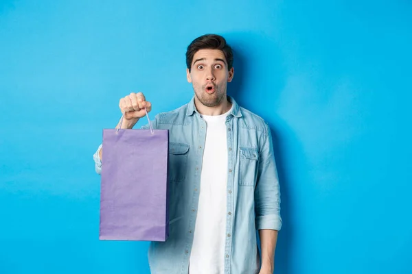 Concepto de compras, vacaciones y estilo de vida. Guapo sorprendido chico sosteniendo bolsa de papel de la tienda y mirando sorprendido, de pie sobre fondo azul —  Fotos de Stock