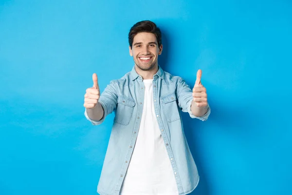 Sorrindo homem confiante mostrando polegares para cima, garantir a qualidade, aprovar algo bom, de pé contra fundo azul — Fotografia de Stock