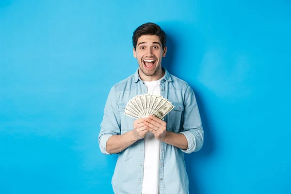Hombre atractivo emocionado y sorprendido, sosteniendo el premio del dinero y sonriendo sorprendido, de pie sobre fondo azul — Foto de Stock