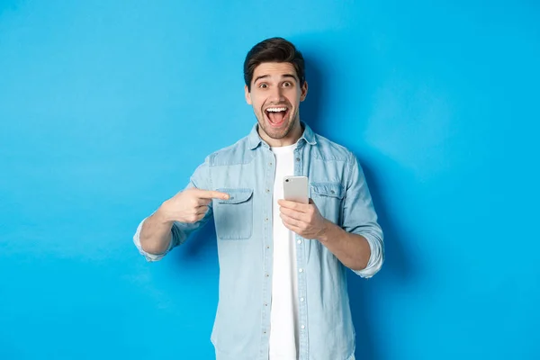 Concept of online shopping, applications and technology. Happy smiling man pointing finger at phone, standing amazed against blue background — Stock Photo, Image