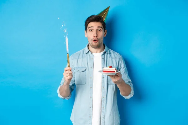 Jovem feliz comemorando aniversário em chapéu de festa, segurando bolo b-day e sorrindo, de pé sobre fundo azul — Fotografia de Stock