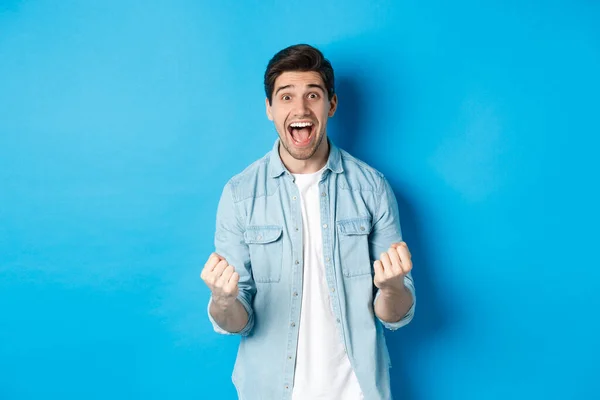 Entusiasmado homem bonito ganhando, sentindo-se sortudo e alegre, comemorando a vitória e gritando sim, de pé contra o fundo azul — Fotografia de Stock