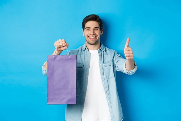 Concepto de compras, vacaciones y estilo de vida. Hombre sonriente satisfecho sosteniendo bolsa de papel, mostrando el pulgar hacia arriba y recomendando tienda, de pie sobre fondo azul —  Fotos de Stock