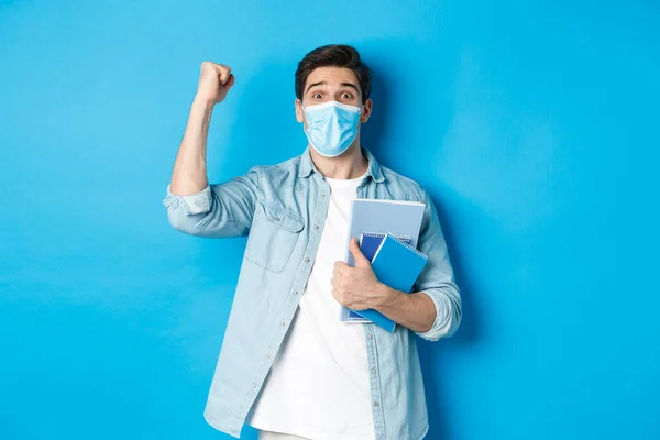 Educação, covid-19 e distanciamento social. Estudante masculino animado em máscara médica triunfando, levantando punho e segurando cadernos, de pé sobre fundo azul — Fotografia de Stock