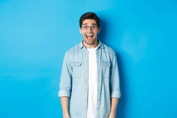Atractivo hombre feliz en gafas mirando sorprendido, mirando publicidad, de pie sobre fondo azul — Foto de Stock