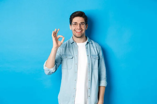 Atractivo joven con gafas y ropa casual, mostrando buena señal de aprobación, como algo, de pie sobre fondo azul — Foto de Stock