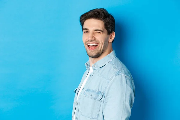 Gelukkige en zelfverzekerde man hoofd draaien op de camera, knipogen en glimlachen, staande over blauwe achtergrond — Stockfoto