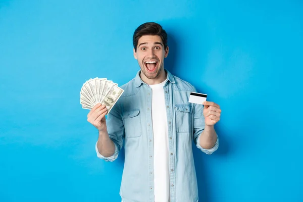 Hombre atractivo feliz mirando sorprendido, mostrando efectivo y tarjeta de crédito, concepto de bancos, crédito y finanzas. Fondo estudio azul — Foto de Stock
