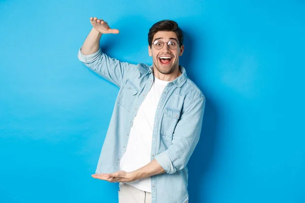 Hombre feliz en gafas mostrando objetos de gran tamaño, dando forma a una caja grande, de pie sobre fondo azul y sonriendo — Foto de Stock