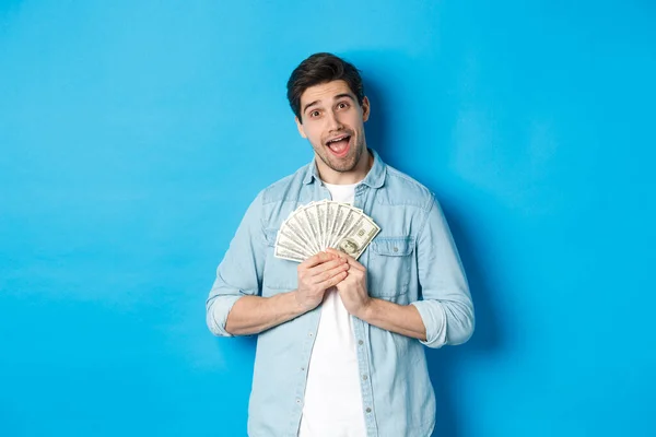 Sonriente chico codicioso abrazando el dinero y sonriendo, no dispuesto a compartir, de pie sobre fondo azul — Foto de Stock