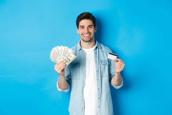 Joven hombre sonriente mostrando dólares en efectivo y tarjeta de crédito, de pie sobre fondo azul — Foto de Stock