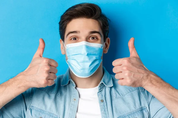 Concept of covid-19, pandemic and quarantine. Close-up of cheerful young man in medical mask smiling, showing thumbs up in approval, like and agree, standing over blue background — Stock Photo, Image