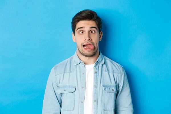 Close-up de jovem fazendo expressões engraçadas, mostrando a língua e olhando para a câmera, de pé sobre o fundo azul — Fotografia de Stock