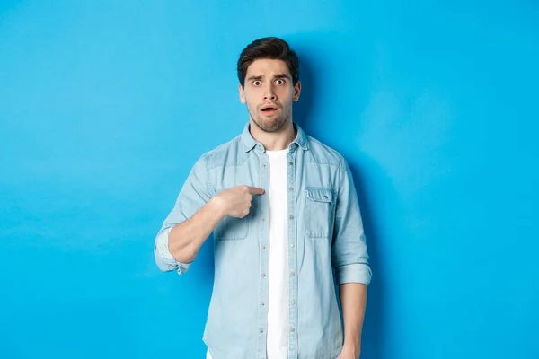 Nervous man pointing at himself and looking confused, standing in casual clothes over blue background