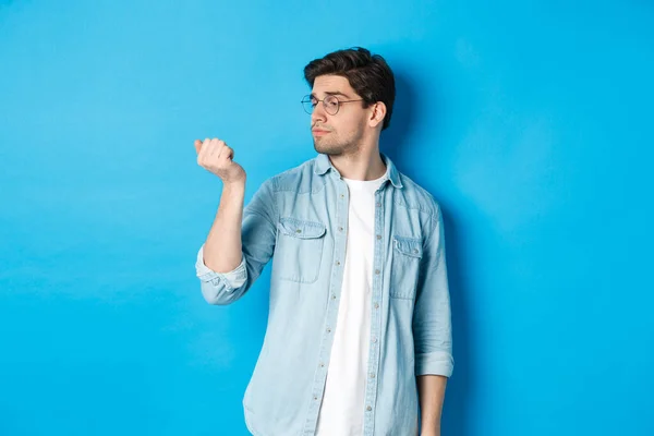 Jonge knappe man met een bril die naar zijn nagels kijkt, manicure controleert, over een blauwe achtergrond staat — Stockfoto