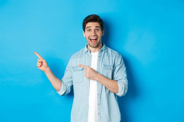 Opgewonden knappe 25s man met baard, wijzende vingers links en lachend verbaasd, staande tegen een blauwe achtergrond — Stockfoto