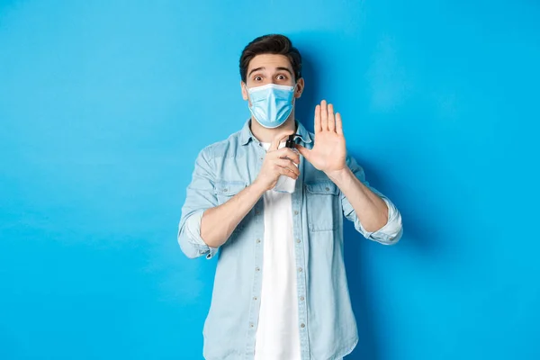 Έννοια του covid-19, πανδημία και κοινωνική αποστασιοποίηση. Χαρούμενος τύπος με ιατρική μάσκα που δείχνει πώς να απολυμαίνουν τα χέρια με απολυμαντικό, χρησιμοποιώντας αντισηπτικό, αποτρέποντας την εξάπλωση του ιού, μπλε φόντο — Φωτογραφία Αρχείου