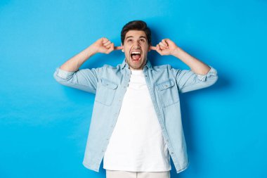 Sinirli adam, gürültücü komşuların kulaklarını kapatır, yukarı bakar ve bağırır, yüksek sesli müzik duyar, mavi arka planda durur.