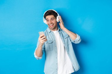 Kulaklıkla müzik dinleyen ve akıllı telefondan mesaj okuyan mutlu bir adam. Heyecanlı bir şekilde gülümsüyor, mavi arka planda duruyor.