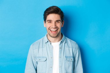 Yakışıklı genç adam gülüyor, günlük kıyafetler giyiyor, mavi arka planda dikiliyor.