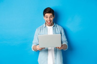 Şaşırmış ve mutlu adamın görüntüsü internetteki özel teklife tepki veriyor, dizüstü bilgisayara heyecanlı bakıyor, mavi arka planda duruyor.