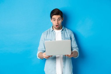Etkileyici beyaz adam şaşkınlıkla dizüstü bilgisayara bakıyor, internetten tanıtıma bakıyor, mavi arka planda duruyor.
