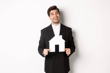 Evini arayan takım elbiseli yakışıklı bir adamın portresi. Elinde kağıt bir ev, sağ üst köşedeki hayallere bakıyor. Beyaz arka planda duruyor.